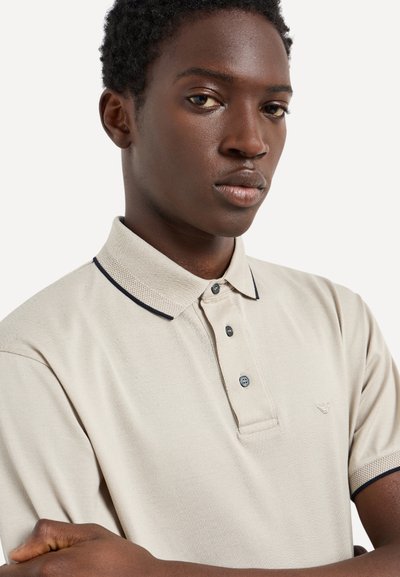 Camisa polo beige con un tejido texturizado, que presenta un cuello con solapa, acentos en azul oscuro y tres botones. Logo bordado en el pecho.