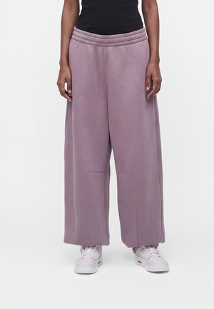 Personne portant un pantalon de survêtement mauve à jambes larges, un haut noir et des baskets rose clair debout sur un fond blanc.
