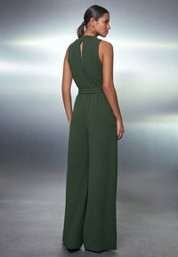 Grön jumpsuit med hög krage, öppen rygg och vida ben. Tyget har en slät textur; inkluderar ett matchande bälte.