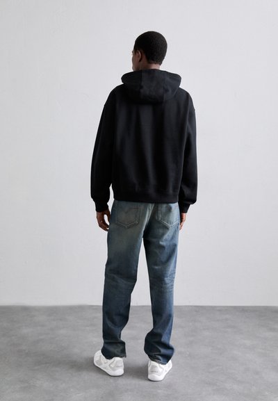 Sudadera negra con capucha, corte holgado, material de algodón. Combinada con jeans de mezclilla azul, acabado desgastado, pierna recta. Zapatillas blancas, con detalles texturizados.