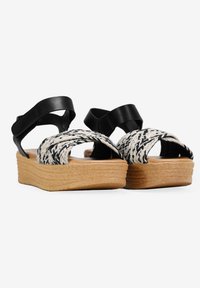 Sandalias de plataforma con tiras de cuero negro y parte superior trenzada en blanco y negro, suela marrón y diseño texturizado, que elevan el estilo y la comodidad.
