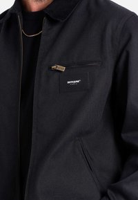 Giacca nera in cotone con tasca sul petto con zip, colletto semplice e etichetta con logo sulla tasca. Tessuto liscio con una vestibilità su misura.