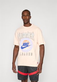 Lyserød bomulds t-shirt med sort tekst, med et centralt blåt og orange cirkulært Nike-logo. Båret med sorte shorts med røde striber.