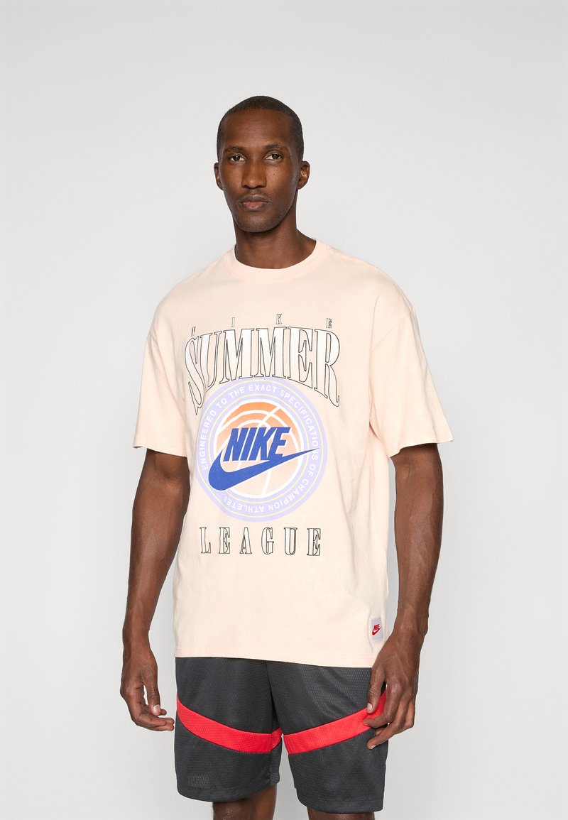 Lyserød bomulds t-shirt med sort tekst, med et centralt blåt og orange cirkulært Nike-logo. Båret med sorte shorts med røde striber.