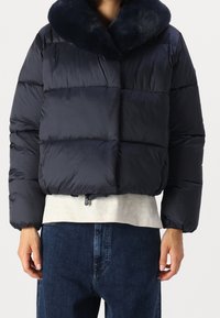 Marinblå dunjacka med mjuk krage, boxig design, sydda sektioner och dragsko i fållen. Kombinerad med en ljusgrå skjorta och denim.