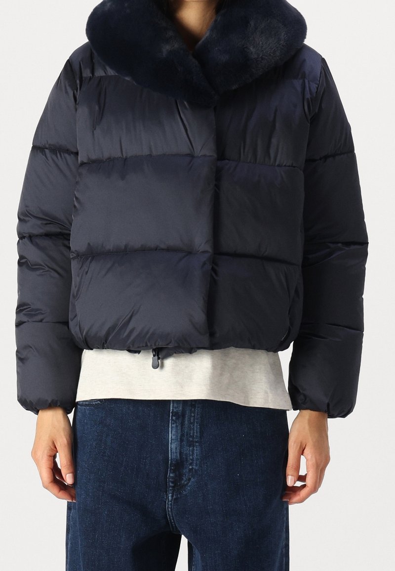 Marinblå dunjacka med mjuk krage, boxig design, sydda sektioner och dragsko i fållen. Kombinerad med en ljusgrå skjorta och denim.