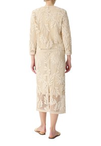 Kremowy zestaw szydełkowy z kwiatowymi wzorami. Kurtka ma rękawy o długości trzech czwartych, a spódnica ma rozcięcie z tyłu.