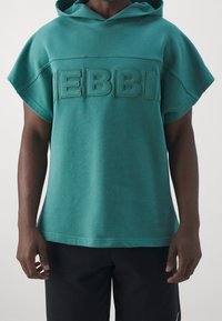 Tealfärgad hooded sweatshirt med korta ärmar, prydd med upphöjd text "DEBBI" på framsidan. Slät textur, avslappnad design.