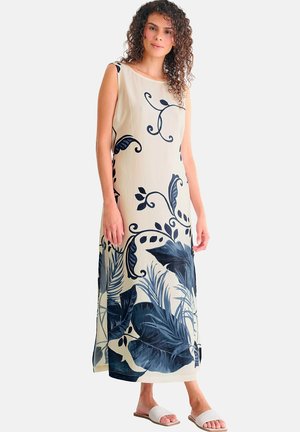 Femme portant une robe maxi beige sans manches avec de grands motifs floraux et de feuilles bleu foncé, associée à des sandales blanches à enfiler.
