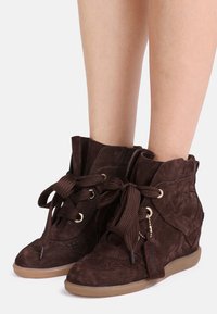 Καφέ σουέτ αθλητικά παπούτσια με σφήνα, στρογγυλερή μύτη, διάτρητες λεπτομέρειες, κορδόνια μπροστά και υφασμένο λαστιχένιο πέλμα. Διαθέτουν χρυσούς κρίκους για τα κορδόνια.