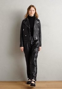 Black leather motorcycle jacket with zippers, paired with black leather pants and black shoes. Model stands on a wooden floor against a light wall.