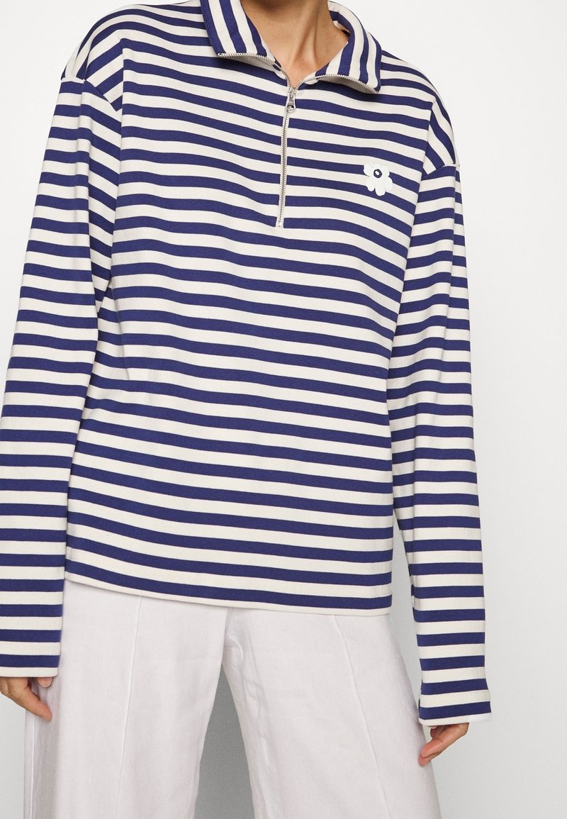 Randig långärmad topp i marinblå och krämfärg, med halvdragkedja i kragen och en blommig accent på bröstet. Slät tygstruktur.