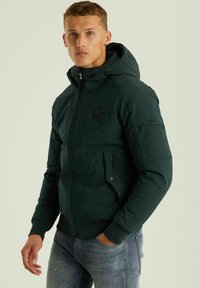 Temno zelena podložena jakna s kapuco, z zadrgo, dvema sprednjima žepoma in majhnim logotipom na prsih. Mehka tekstura in prilegajoč dizajn.
