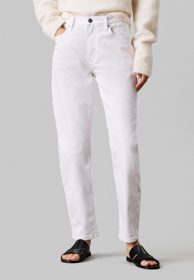 Białe dżinsy z prostym krojem nogawki, wysokim stanem, pięcioma kieszeniami i subtelną teksturą. W połączeniu z czarnymi klapkami.