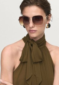 Oversized brown gradient sunglasses with gold-tone frame and geometric shape. Model wears olive green halter top with a knotted detail.