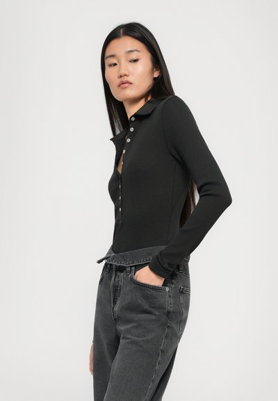 Young woman with long dark hair wears a black buttoned long-sleeve top tucked into high-waisted loose gray jeans, hand in pocket.