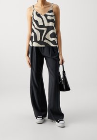 Femme portant un haut sans manches à imprimé abstrait noir et blanc, un pantalon noir fluide, des baskets noir et blanc, tenant un petit sac à main noir.