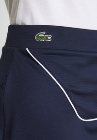 Marinblå skort med en slät textur, utrustad med vit sidopiping och en grön broderad krokodillogga på midjebandet.