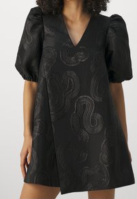 Schwarzes Kleid mit V-Ausschnitt und Puffärmeln. Verfügt über ein strukturiertes Wirbelmuster in metallischen Fäden, glattes Gewebe und eine A-Linien-Silhouette.