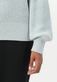 Svetlo modra pletena jopica z rebrastimi manšetami in robom. Tekstura je mehka in ima ohlapno prileganje, kar poudarja sproščen dizajn.