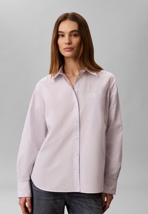 Frau trägt ein hellviolettes gestreiftes, langärmliges Hemd mit Knöpfen und dunkle Jeans, steht vor einem grauen Hintergrund.