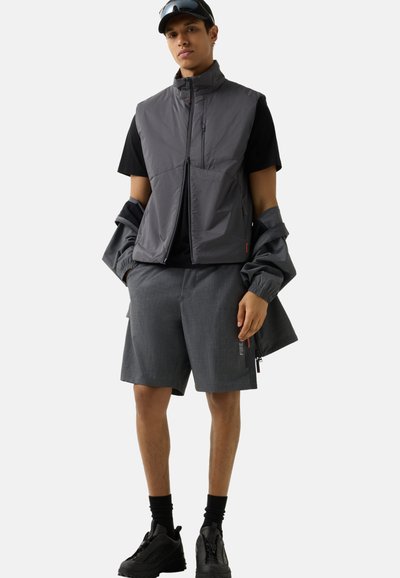 Jonge man met een grijze mouwloze vest, bijpassende korte broek, zwart T-shirt, zwarte sokken, zwarte sneakers en zonnebril op het hoofd tegen een effen achtergrond.