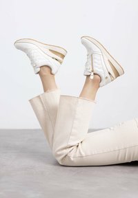 Zapatillas acolchadas blancas con un acento dorado y suela beige, que cuentan con un zipper lateral y cordones, combinadas con pantalones de pierna ancha color beige claro.