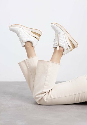Zapatillas acolchadas blancas con un acento dorado y suela beige, que cuentan con un zipper lateral y cordones, combinadas con pantalones de pierna ancha color beige claro.
