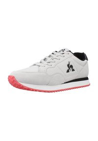 Zapatilla deportiva blanca con suela rosa, acentos negros y parte superior texturizada. Presenta una puntera redondeada y un sistema de cordones en la parte delantera con detalles del logo.