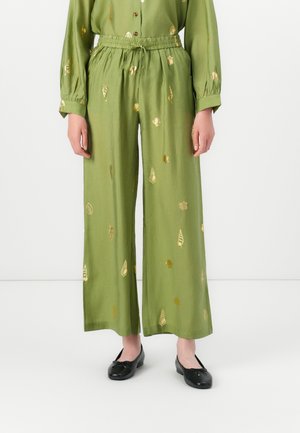 Pantaloni larghi verdi con motivi a conchiglia dorati, cintura elastica, indossati con ballerine nere su uno sfondo bianco.