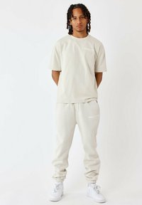 Camiseta de algodón beige y joggers a juego, con un pequeño logo blanco en el pecho y la pierna, complementados con zapatillas blancas.