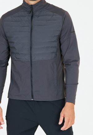 Homme portant une veste zippée gris foncé avec poitrine matelassée et manches ainsi que bas devant en tissu lisse, debout sur fond blanc.