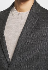 Grauer strukturierter Blazer mit Reverskragen, kombiniert mit einem hellbeigen gerippten Pullover, der ein geschichtetes, strukturiertes Design zeigt.