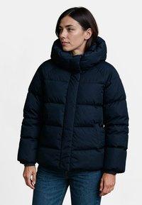 Marine blauwe gewatteerde jas met een hoge kraag, voorzien van horizontaal gestikte secties, een verborgen rits en elastische manchetten. Draagt over spijkerbroeken.