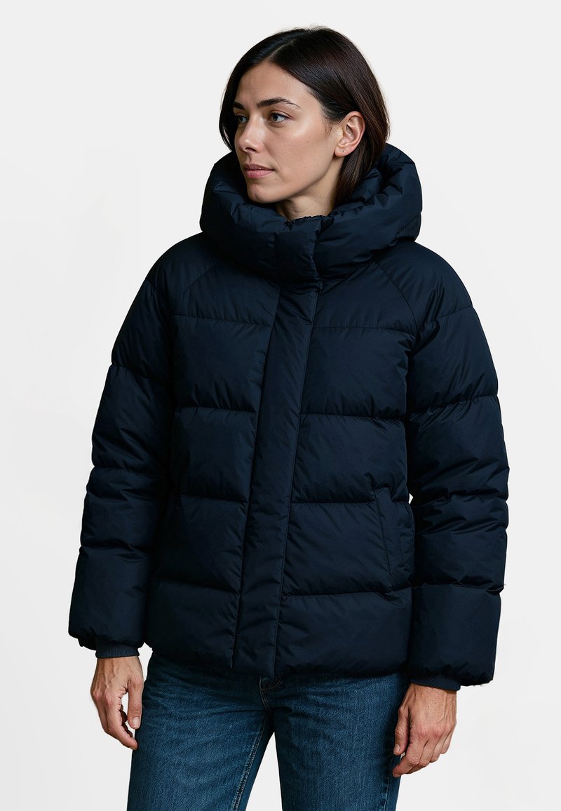 Marine blauwe gewatteerde jas met een hoge kraag, voorzien van horizontaal gestikte secties, een verborgen rits en elastische manchetten. Draagt over spijkerbroeken.