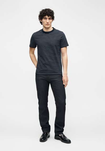 Άνδρας που στέκεται φορώντας μαύρο και άσπρο ριγέ κοντομάνικο μπλουζάκι, σκούρο τζιν και μαύρα παπούτσια σε απλό φωτεινό φόντο.