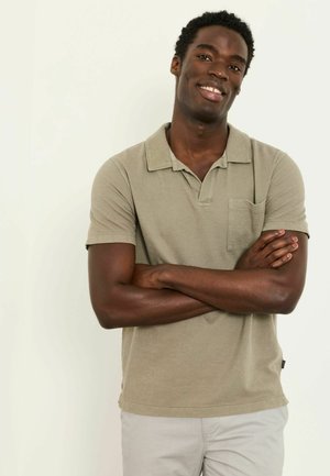 Naeratav noormees, käed rüpes, kandes beeži poloshirt ja heledaid pükse, seisab ühevärvilise valge tausta ees.