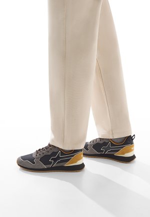 Sportliche Sneakers mit einem schwarzen Mesh-Obermaterial und grauen sowie goldenen Akzenten, strukturiertem Profil und unterstützendem Design; kombiniert mit beigen Hosen.
