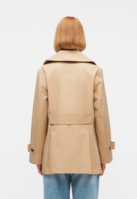 Γυναίκα με ίσια, μακριά μέχρι τους ώμους μαλλιά, που φοράει ένα μπεζ trench coat και μπλε τζιν, εμφανίζεται από πίσω με φόντο λευκό.