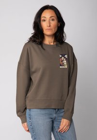 Frau mit dunklem Haar, die einen braunen Sweatshirt mit einem Tarotkarten-Design "Die Liebenden" und hellblauen Jeans trägt, steht vor einem schlichten Hintergrund.