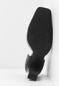 Chaussure à talon noir avec une finition brillante ; bout carré et semelle texturée. Dispose d'un talon bloc de hauteur intermédiaire et d'un intérieur lisse.