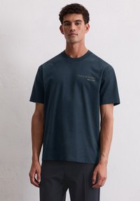 Dunkelblaues Baumwoll-T-Shirt mit kurzen Ärmeln, Rundhalsausschnitt und kleinem goldenen Textdetail auf der Vorderseite. Glatte Textur, entspannter Schnitt.