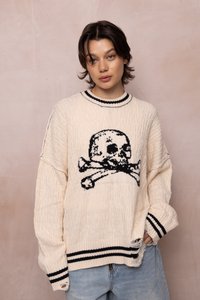 Joven con cabello corto y oscuro, vistiendo un suéter de punto color crema con un diseño de calavera y tibias cruzadas en negro, acompañado de jeans azul claro, frente a una pared rosa.