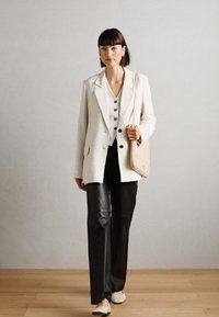 Blazer blanc à rayures fines sur un haut noir, pantalon en cuir noir et chaussures beiges ; portant un sac à bandoulière beige. Textures lisses et design soigné.
