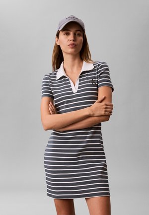 Kvinne som har på seg en grå og hvit stripet polo-kjole og matchende grå caps, står med armene i kors mot en ensfarget bakgrunn.