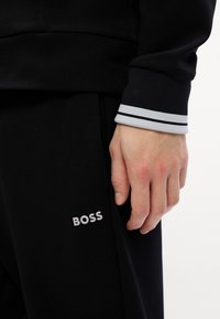 Melna sporta jaka ar svītrām pie piedurknēm, kurai ir smalka pelēka logo "BOSS" uz biksēm. Mīksts audums, pieguļošs dizains.