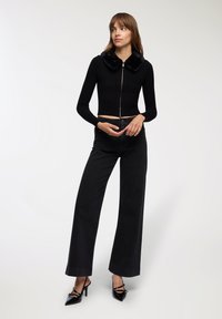 Kobieta stoi w czarnej, prążkowanej bluzie zapinanej na zamek z kołnierzem z sztucznego futra, szerokich czarnych dżinsach oraz czarnych szpilkach w kształcie czubka.