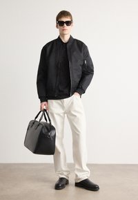 Schwarze Bomberjacke mit einem Reißverschluss vorne und einer Taschen an den Ärmeln, kombiniert mit einem schwarzen T-Shirt, cremefarbenen Jeans sowie einer schwarzen Lederhandtasche und Schuhen.