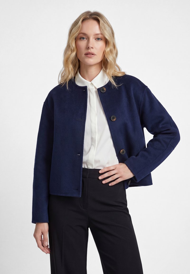Femme blonde aux cheveux ondulés porte une veste bleu marine boutonnée, une chemise blanche à col et un pantalon noir, posant avec une main sur la hanche.