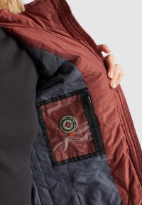 Burgunderrød quilted jakke med marineblå inderside, der har en gennemsigtig lomme og rund logo-patch. Glat, slidstærk stofstruktur synlig.
